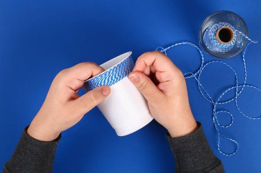 DIY kalem tutucu plastik bardak ekşi krema, yoğurt iplik mavi arka plan sarılmış. Dekorasyon çiçek sıcak tutkal, dekoratif yapışkan bant. Hediye fikri. Adım adım. Üstten görünüm. Süreci çocuk çocuk zanaat