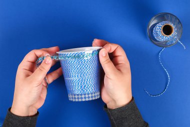 DIY kalem tutucu plastik bardak ekşi krema, yoğurt iplik mavi arka plan sarılmış. Dekorasyon çiçek sıcak tutkal, dekoratif yapışkan bant. Hediye fikri. Adım adım. Üstten görünüm. Süreci çocuk çocuk zanaat
