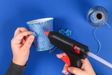 DIY kalem tutucu plastik bardak ekşi krema, yoğurt iplik mavi arka plan sarılmış. Dekorasyon çiçek sıcak tutkal, dekoratif yapışkan bant. Hediye fikri. Adım adım. Üstten görünüm. Süreci çocuk çocuk zanaat