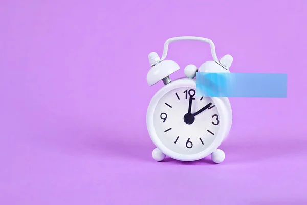 Reloj despertador blanco violeta fondo pastel. Nota pegajosa en blanco en el reloj. Copia espacial. Concepto mínimo . —  Fotos de Stock