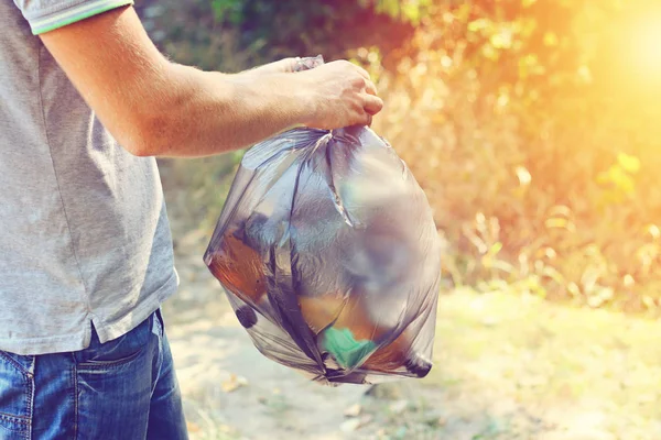 Hand hält gegen Wald voller Müll schwarze Plastiktüte, Sommertag, Müllabfuhr in der Natur. — Stockfoto