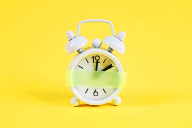 Beyaz çalar saat sarı pastel arka plan. Saat üzerinde boş yapışkan not. Uzay kopyası. Minimal konsept.