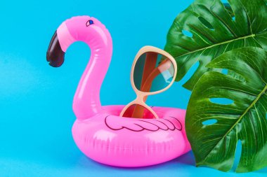 Mavi arka planda pembe şişirilebilir flamingo, güneş gözlüğü ve canavar yapraklı..