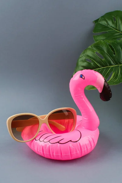 Rosa uppblåsbar flamingo på grå bakgrund med solglasögon och monstera blad. — Stockfoto