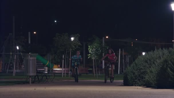 Duas crianças andando de bicicleta thier em um parque à noite — Vídeo de Stock