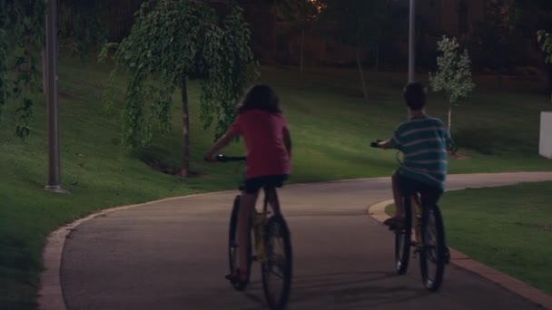 Duas crianças andando de bicicleta thier em um parque à noite — Vídeo de Stock