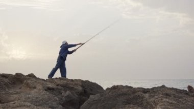 Deniz tarafında kayaların üzerinde duran ve günbatımı karşı Balık tutma eski balıkçı