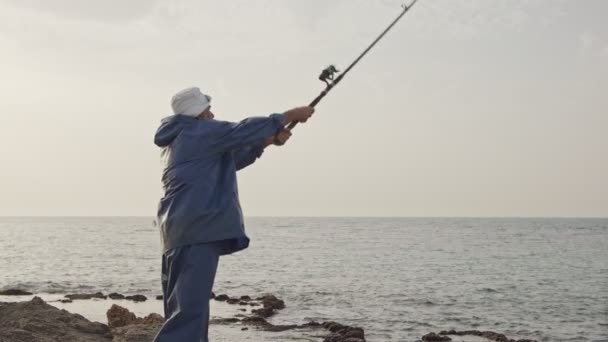 Gamla fiskare står på havet sida stenar och fiske mot solnedgången — Stockvideo