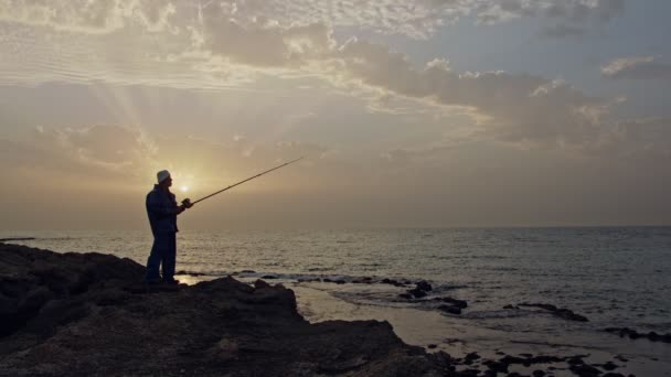 Γέρος ψαράς στέκεται πάνω σε βράχια πλάι στη θάλασσα και το ψάρεμα κατά το ηλιοβασίλεμα — Αρχείο Βίντεο