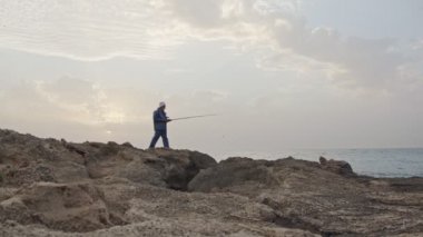 Deniz tarafında kayaların üzerinde duran ve günbatımı karşı Balık tutma eski balıkçı