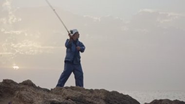 Deniz tarafında kayaların üzerinde duran ve günbatımı karşı Balık tutma eski balıkçı
