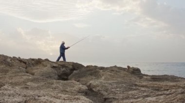Deniz tarafında kayaların üzerinde duran ve günbatımı karşı Balık tutma eski balıkçı
