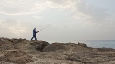 Deniz tarafında kayaların üzerinde duran ve günbatımı karşı Balık tutma eski balıkçı