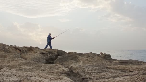 Velho pescador de pé no lado do mar rochas e pesca contra o pôr do sol — Vídeo de Stock