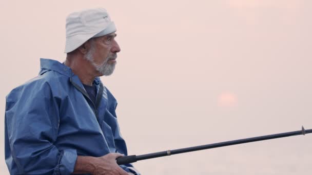 Vieux pêcheur debout sur les rochers du bord de la mer et la pêche contre le coucher du soleil — Video