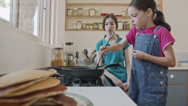 Kaksi nuorta tyttöä valmistelee pannukakkuja keittiössä paistinpannulla — kuvapankkivideo