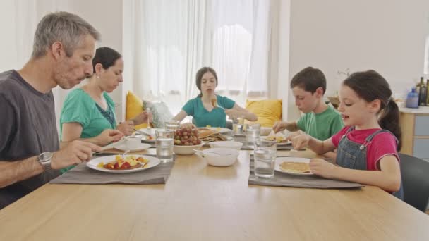 Familia de cinco personas sentadas a la mesa comiendo panqueques y frutas para desayunar — Vídeos de Stock