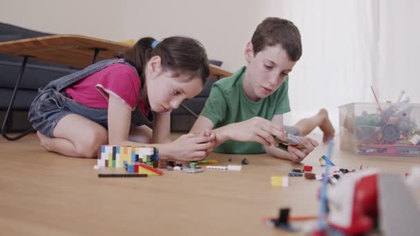 Lány és egy fiú játszanak és építkeznek játéktéglákkal a nappali padlóján — Stock videók