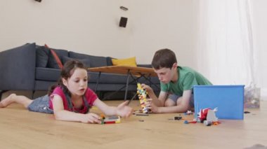 Bir kız ve bir oğlan oturma odasının zemininde oyuncak tuğlalarla oynayıp inşa ediyorlar.