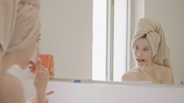 Genç kız banyo aynasının önünde dudak kremi ve dudak parlatıcısı sürüyor.