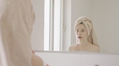 Genç kız banyo aynasının önünde dudak kremi ve dudak parlatıcısı sürüyor.