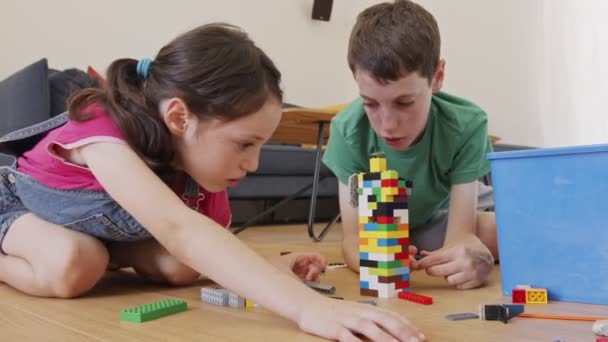 Lány és egy fiú játszanak és építkeznek játéktéglákkal a nappali padlóján — Stock videók