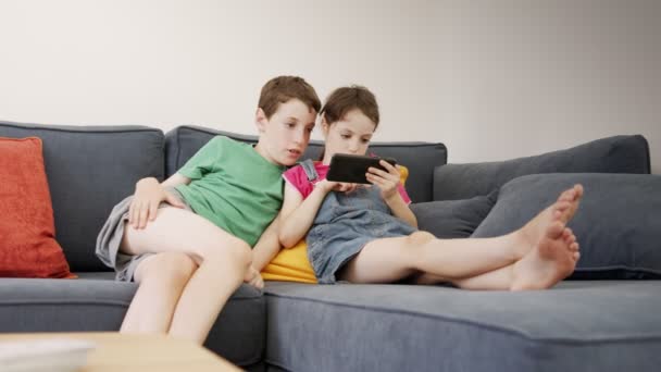 Két gyerek ül otthon egy nagy kanapén, mobiltelefont használva. — Stock videók