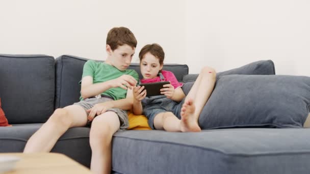 Twee kinderen die thuis op een grote bank zitten, met een mobiele telefoon — Stockvideo