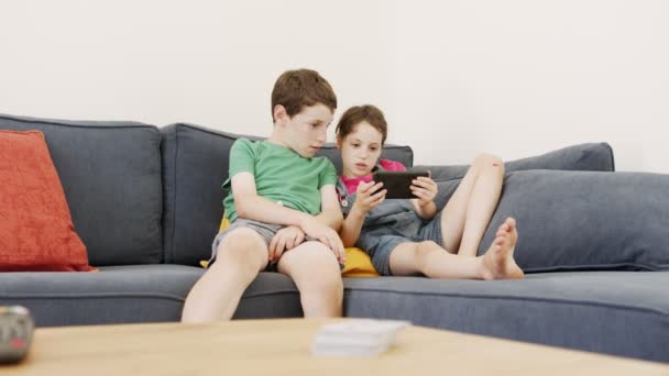 Dos niños sentados en un sofá grande en casa, usando un teléfono móvil — Vídeos de Stock