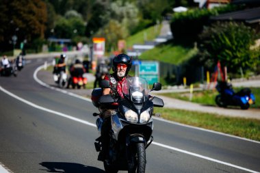 Faaker See, Avusturya - 01 Eylül 2025: Avrupa Bisiklet Haftası boyunca motosiklet süren beyaz sakallı erkek motosikletçi, çevresi manzaralı yollar ve motosikletçilerle çevrili