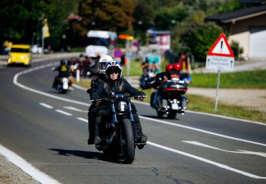 Faaker See, Avusturya - 01 Eylül 2025: Deri ceketli bisikletçi kadın Avrupa Bisiklet Haftası sırasında motosikletini sürüyor, çevresi motosikletli arkadaşlarıyla manzaralı bir yolda