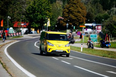 Faaker See, Avusturya - 01 Eylül 2025: Sarı teslimat aracı Avrupa Bisiklet Haftası boyunca arka planda motosikletçiler ve yeşilliklerle birlikte manzaralı bir yolda ilerliyor