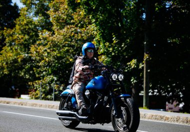 Faaker, Avusturya - 01 Eylül 2025: Mavi kasklı erkek sürücü canlı ağaçlarla çevrili özel mavi bir motosikletle geziyor