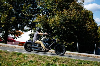 Faaker See, Avusturya - 01 Eylül 2025: Erkek motorcu ağaçlar ve festival atmosferi olan manzaralı bir yolda siyah bir Harley-Davidson motosikleti sürüyor