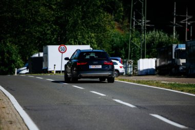 Faaker See, Avusturya - 01 Eylül 2025: Siyah Audi aracı yeşil alan ve görünür hız limiti işareti ile çevrili kırsal bir yolda ilerliyor