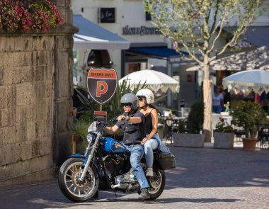 Villach, Avusturya - Eylül 04, 2025: Motosikletli ikili, kafeler ve çiçeklerle dolu güzel bir caddede klasik bir motosikletin keyfini çıkarıyor