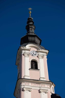 Villach, Avusturya - Eylül 04, 2025: Tarihi kilise kulesinde, canlı mavi gökyüzüne karşı dizilmiş, mimari zarafeti vurgulayan bir saat ve altın haç yer alıyor