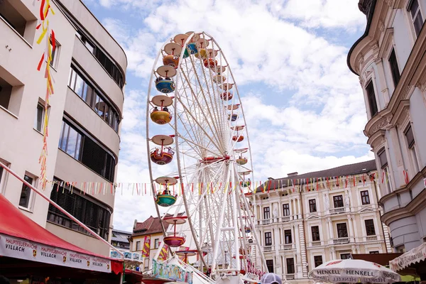 Villach, Avusturya - 30 Temmuz 2025: Villacher Kirchtag festivalinde büyüleyici mimari ve renkli dekorasyonlarla çerçevelenmiş canlı dönme dolap, neşeli bir ortam yaratıyor.