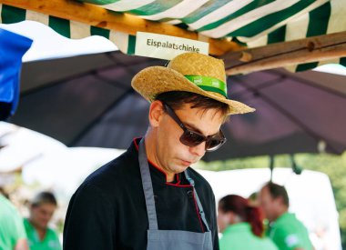 Ossiacher Tauern, Avusturya - 10 Ağustos 2025: Hasır şapkalı ve güneş gözlüklü erkek aşçı, festival ortamlı Rindfleischfest 'te gıda hazırlıklarına odaklandı