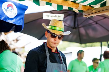 Ossiacher Tauern, Avusturya - 10 Ağustos 2025: Hasır şapkalı ve güneş gözlüklü erkek aşçı festival ortamlarıyla çevrili Rindfleischfest 'te geleneksel yemek hazırlıyor