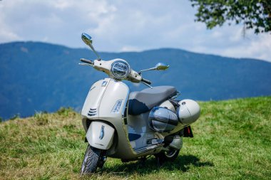 Ossiacher Tauern, Avusturya - 10 Ağustos 2025: Vespa scooter arkasında dağlar olan yeşil çimlerin üzerinde duruyor, parlak tasarımını ve açık hava yaşam tarzını ön plana çıkarıyor
