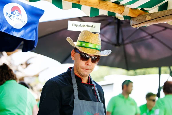 Ossiacher Tauern, Avusturya - 10 Ağustos 2025: Hasır şapkalı ve güneş gözlüklü erkek aşçı festival ortamlarıyla çevrili Rindfleischfest 'te geleneksel yemek hazırlıyor