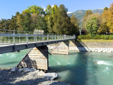 Lienz, Avusturya - 18 Eylül 2025: Çağdaş yaya köprüsü, canlı ağaçlar ve sakin manzarayla çerçevelenmiş, doğanın güzelliğini vurgulayan turkuaz nehri geçiyor