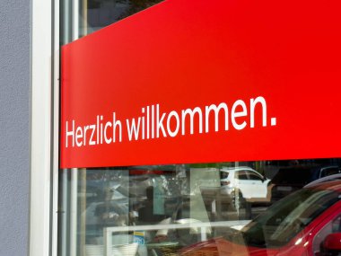Wolfsberg, Avusturya - 19 Eylül 2025: Mağaza vitrininde parlak kırmızı karşılama tabelası, müşterileri Almanca dostça selamlamaya davet ediyor