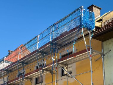 Bleiburg, Avusturya - 19 Eylül 2025: Mavi ağlı iskeleler parlak bir gökyüzüne karşı kurulmuş, bu da bina üzerinde devam eden inşaat çalışmalarını gösteriyor