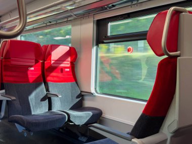 Bleiburg, Avusturya - 19 Eylül 2025: Modern trenin iç mekanında kırmızı ve gri koltuklar sergilenmekte ve güneş ışığı mekanı aydınlatarak seyahat deneyimini artırmaktadır
