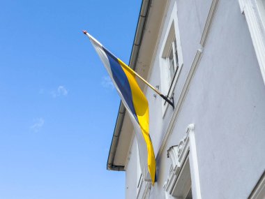 Mavi ve sarı bayrak açık gökyüzüne karşı beyaz binada dalgalanıyor.