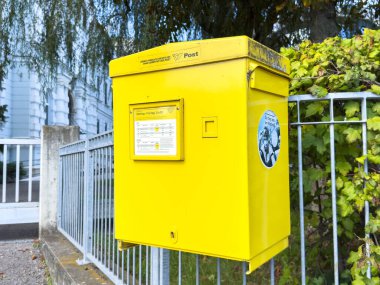 Sankt Veit Ad Glan, Avusturya - 17 Eylül 2025: Parlak sarı posta kutusu, yemyeşil bir çitin yanında, kentsel posta hizmetini temsil eden,