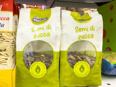 Gemona, İtalya - 24 Eylül 2025: Süpermarket raflarında sergilenen paketlenmiş balkabağı tohumları, canlı renkler ve sağlıklı abur cubur etiketleri için açık ürün