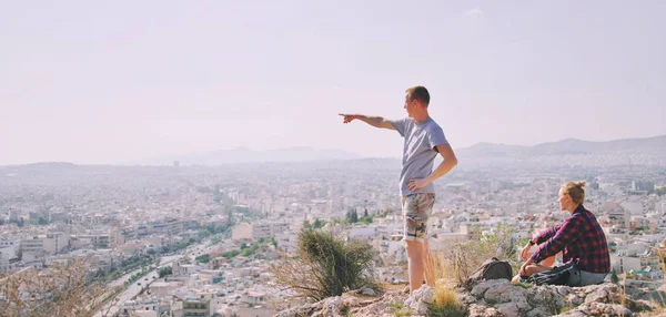 Ζευγάρι Άνδρας και Γυναίκα στην κορυφή του βουνού απολαμβάνοντας το όμορφο τοπίο cityscape Athens Greece στο φόντο. Οι τουρίστες χαλαρώνουν στο λόφο της Αθήνας το καλοκαίρι. Διάσημη Αθήνα της Ευρώπης. ενεργές διακοπές. — Φωτογραφία Αρχείου
